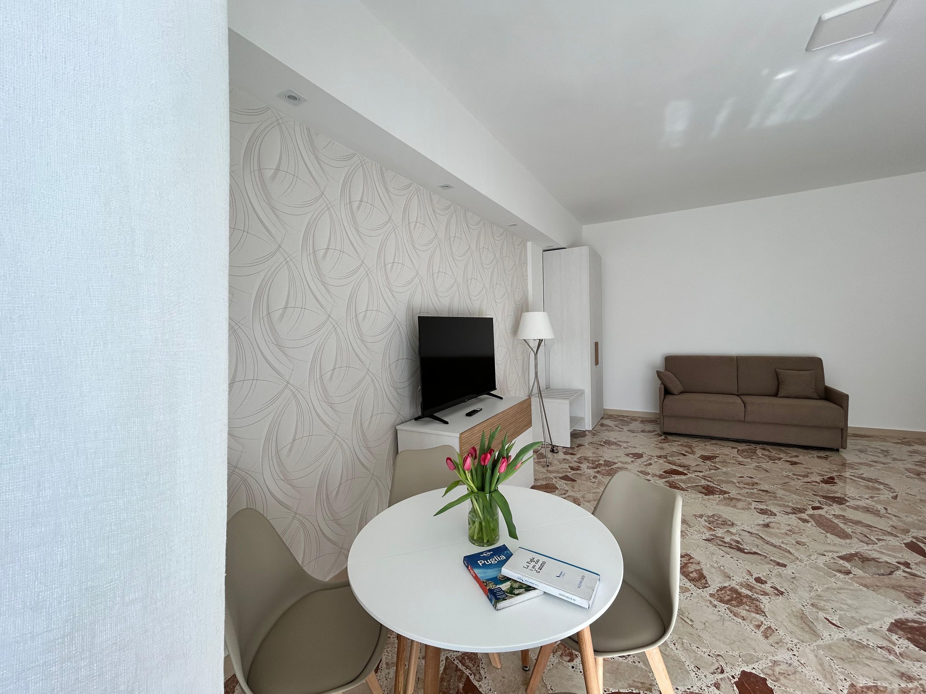 A modern living room with a flat-screen TV, a sofa, a dining table with chairs, and a patterned wall.