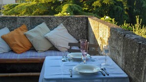 Une table dressée pour deux personnes, avec une nappe bleue, des assiettes blanches, des couverts en argent et des verres roses, placée à côté d’un mur de pierre sur lequel se trouve un banc et des coussins.