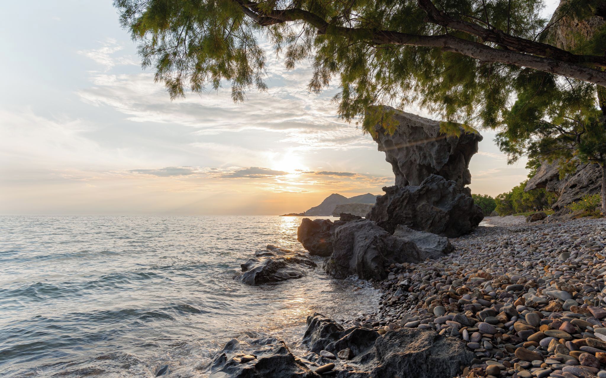 A rocky beach with a large rock formation, calm sea, and a sunset.
