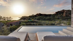 A poolside area with lounge chairs, a view of the sea, and a mountainous landscape.