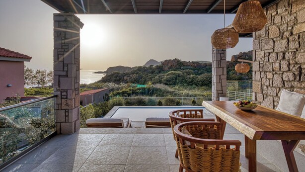 A terrace with a view of the sea, stone walls, wicker furniture, and a wooden table with a bowl of fruit.