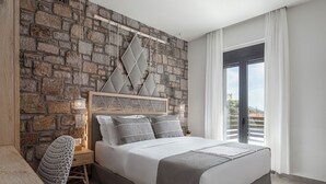 A bedroom with a stone wall, a bed with grey bedding, a wooden nightstand, a woven chair, and a view through a large window.