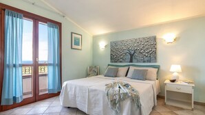 A bedroom with a bed, a nightstand, a wall-mounted lamp, a painting, and a view through a window with curtains.