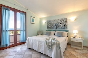 A bedroom with a bed, a nightstand, a wall-mounted lamp, a painting, and a view through a window with curtains.
