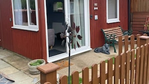 Egy piros színű, fából készült ház, csúszó üvegajtóval, egy kis terasszal, ahol egy pad található, és egy fa kerítéssel.