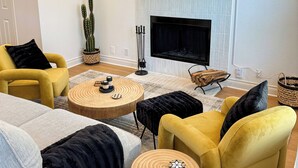 Een woonkamer met een open haard, gele fauteuils, een ronde salontafel en een televisie boven de open haard.