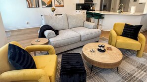 Een moderne zithoek met een bank, gele fauteuils, een ronde salontafel en een keuken op de achtergrond.