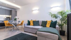 A modern living room with a grey sofa, yellow and green cushions, a dining table with chairs, and a potted plant. - Linford Luxe | Quiet 1-Bed Retreat in Milton Keynes (Linford Wood)