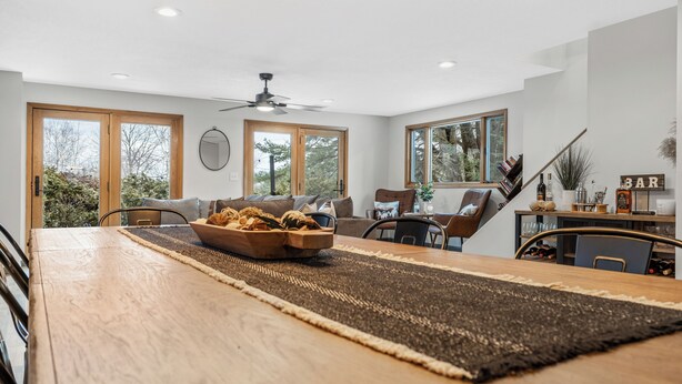 A modern living room with a dining area, large windows, and a ceiling fan.