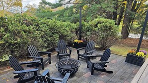 A deck with Adirondack chairs and a fire pit, surrounded by greenery and flower beds.