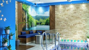 Un espace piscine intérieur moderne, aux couleurs bleu et beige, agrémenté d’une fresque représentant une cascade et d’un coin salon avec des coussins bleus.