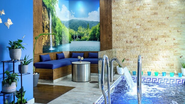 Une salle de spa avec une fresque représentant une cascade, un coin salon, un jacuzzi et un mur en pierre.