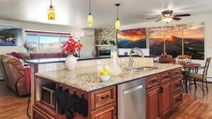 A modern kitchen with granite countertops, stainless steel appliances, and a central island. There are pendant lights, a ceiling fan, and artwork on the walls.