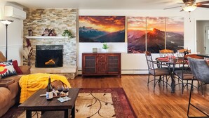 A living room with a fireplace, a dining area with a table and chairs, and a large painting of a sunset over mountains.