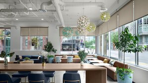 A modern interior with a long wooden table, blue bar stools, a sofa, and large windows offering a view of the street.