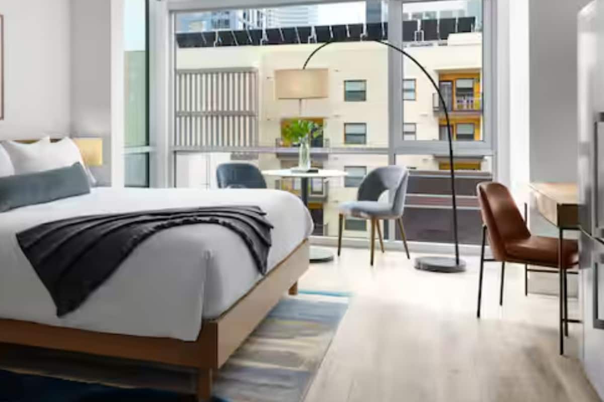 Habitación de hotel moderna con una cama grande, mesita de noche, lámpara y vistas de la ciudad a través de amplios ventanales.