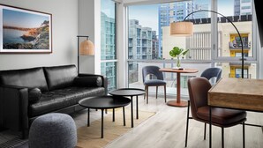 Una sala moderna con un sofá de cuero negro, dos mesas redondas y una mesa de comedor con sillas. Un ventanal ofrece vistas a edificios de la ciudad.