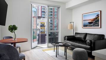 A modern living room with a black leather sofa, a round wooden coffee table, and a grey pouf. A large window offers a view of a building with balconies.