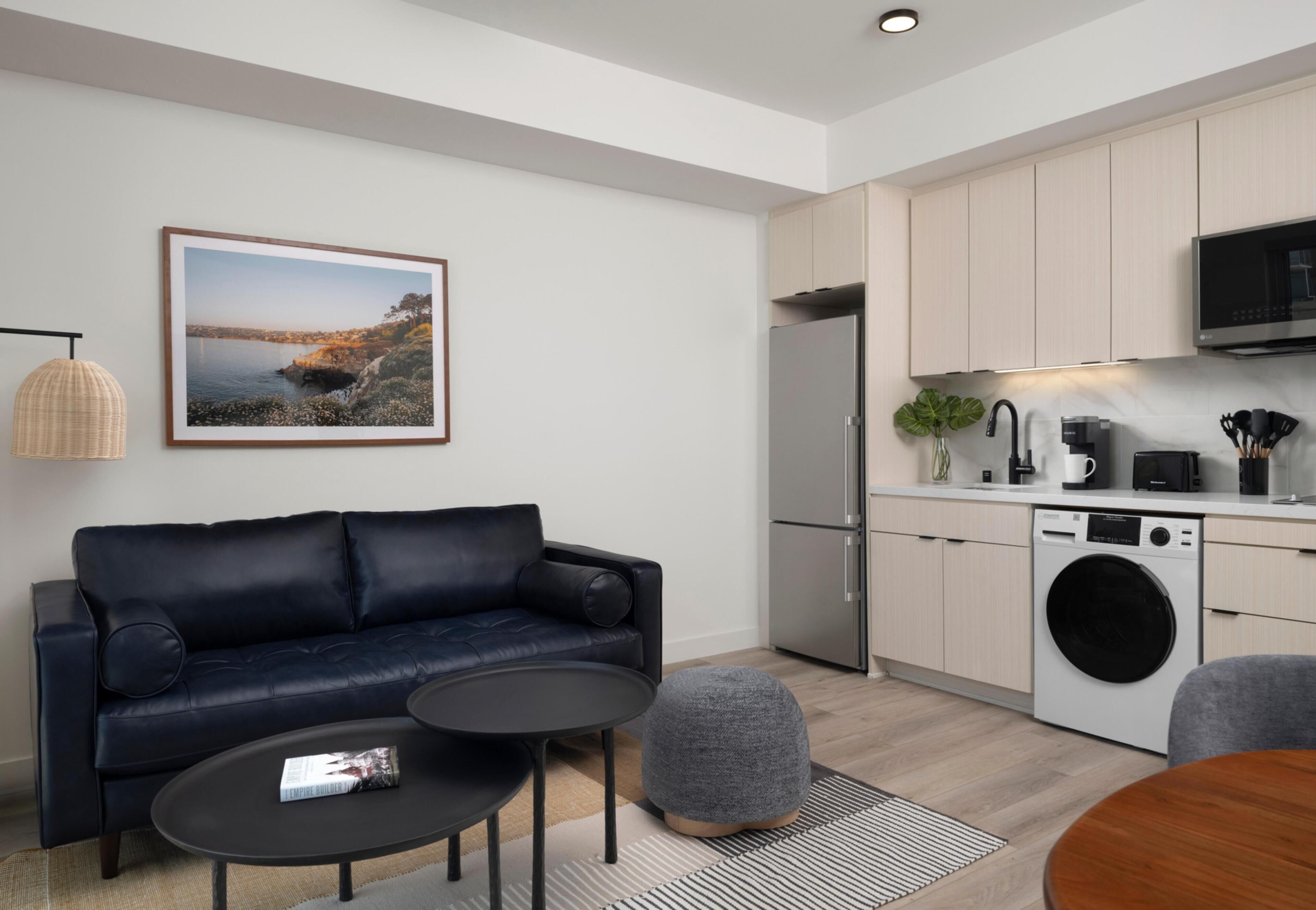 A modern living room with a sofa, coffee table, and ottoman, a kitchen with appliances, and a framed picture on the wall.
