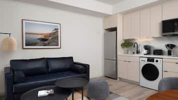 A modern living room with a sofa, coffee table, and ottoman, a kitchen with appliances, and a framed picture on the wall.