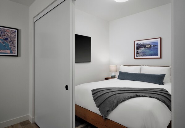Un dormitorio con cama, mesita de noche, lámpara, un cuadro enmarcado en la pared y una puerta.