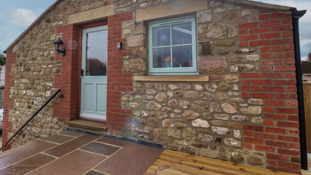 A small stone and brick building with a white door, a window with a light blue frame, and a wooden ramp leading to the entrance. - The Hayloft (Richmond)