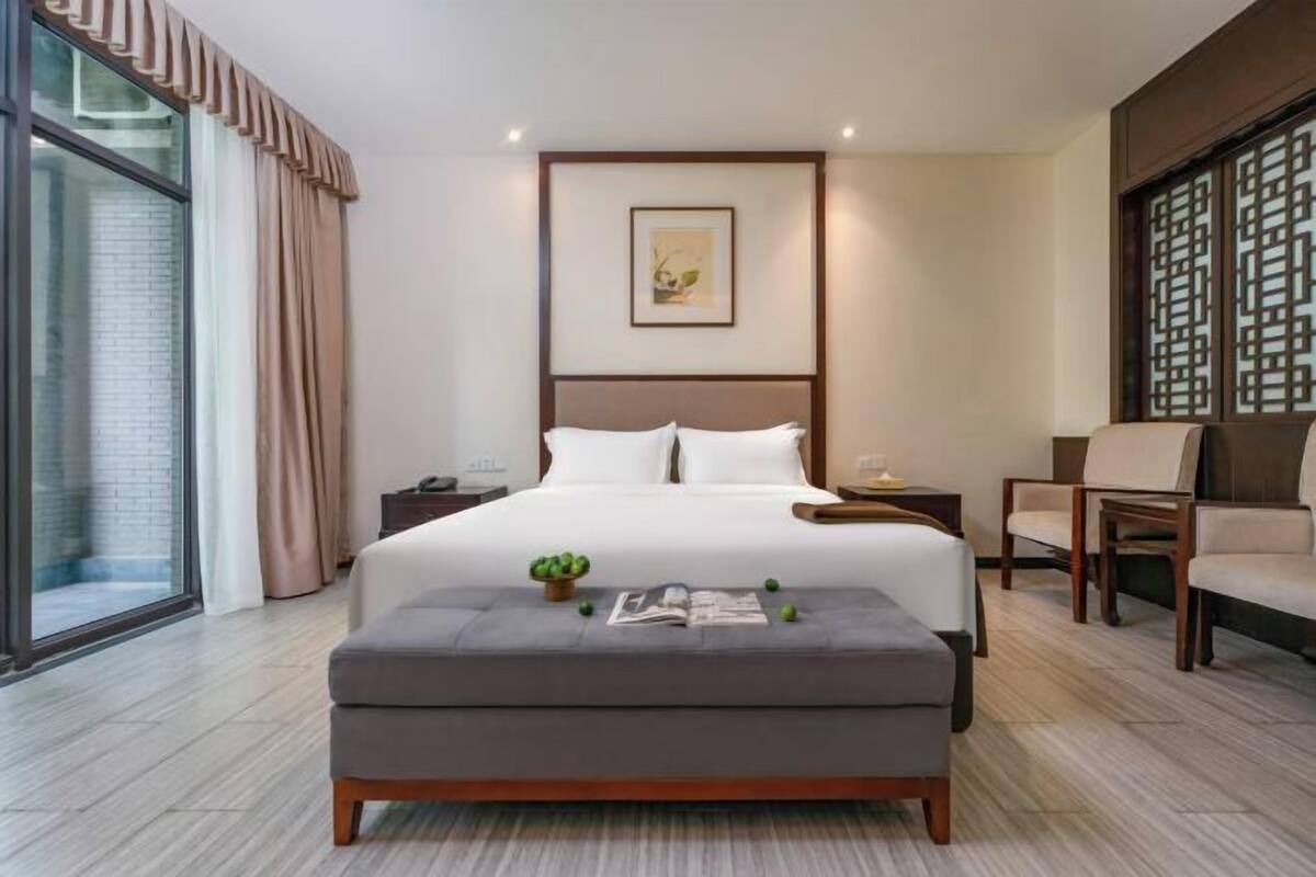 A modern hotel room with a large bed, a bedside table, a reading lamp, a small table with a plant, and a large window with curtains.