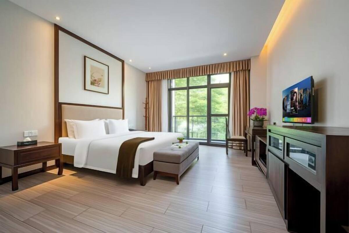 A hotel room with a large bed, a bedside table, a small table with a plant, a view of greenery through the window, and a kitchen area with a microwave.