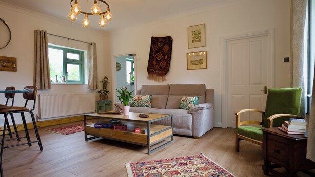 A living room with a sofa, a coffee table, a chair, a bookshelf, a window with curtains, and a hanging light fixture. - Tring Grange Cottage - Sleeps 2 - Onsite Parking (Tring)