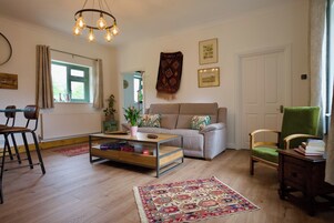 Una sala de estar con un sofá, una mesa de centro, una silla, una estantería, una ventana con cortinas y una lámpara colgante.