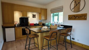 A modern kitchen with wooden cabinets, a central island with bar stools, a built-in oven, and a wall-mounted clock. - Tring Grange Cottage - Sleeps 2 - Onsite Parking (Tring)
