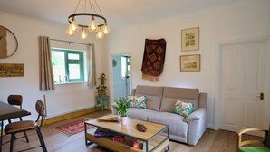 A living room with a sofa, coffee table, and chairs. There is a window with curtains, a plant, and a wall-mounted light fixture. - Tring Grange Cottage - Sleeps 2 - Onsite Parking (Tring)