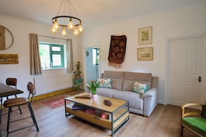 Una sala de estar con sofá, mesa de centro y sillas. Hay una ventana con cortinas, una planta y un aplique de pared.