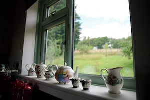 Un alféizar con varios teteras y tazas de cerámica, una taza roja, y vista a un campo verde a través de la ventana.