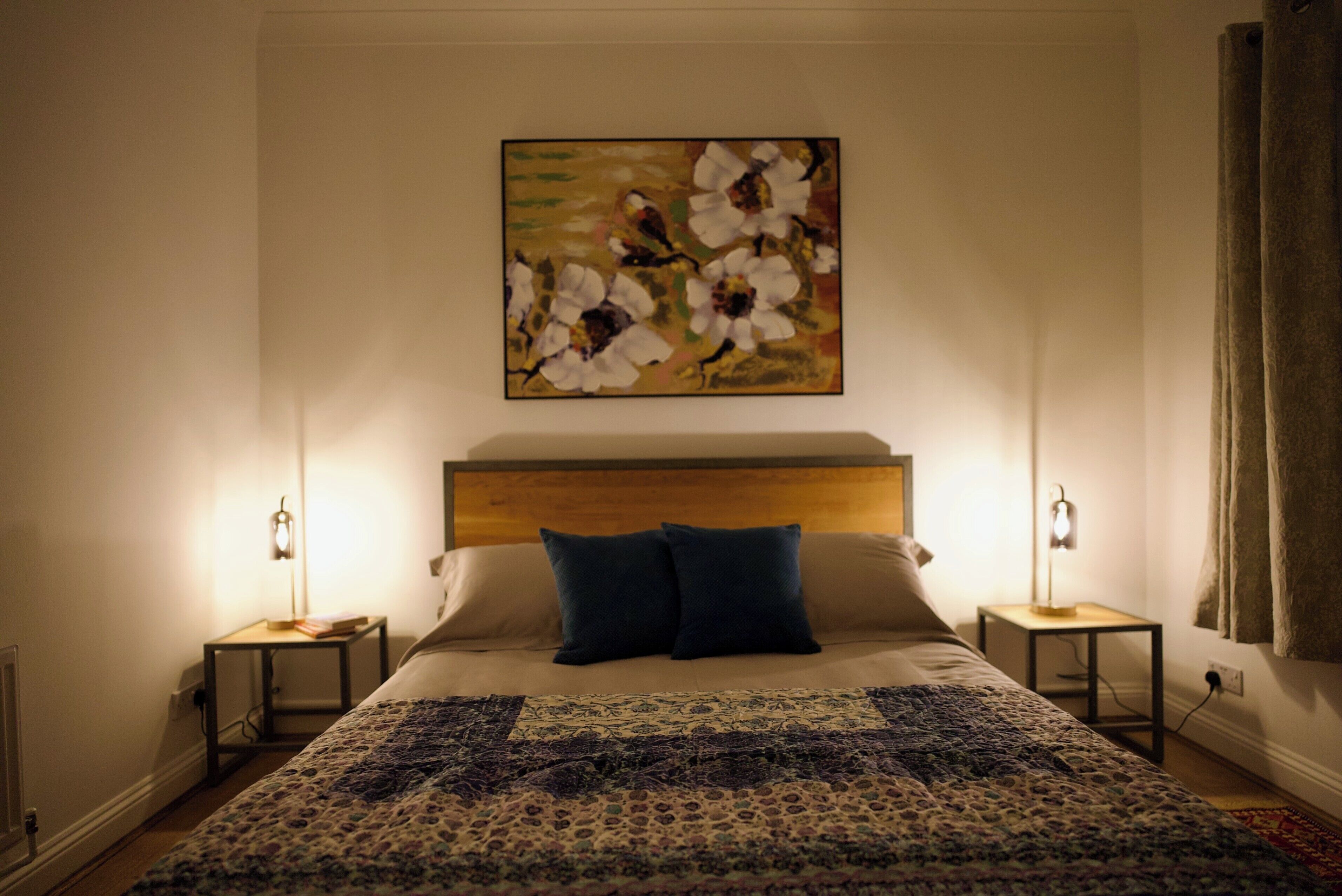 A bedroom with a bed, two bedside tables, a headboard, a large floral painting, and a patterned bedspread.