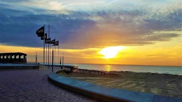 Solnedgangsudsigte fra en strandpromenade med en mole og flag.