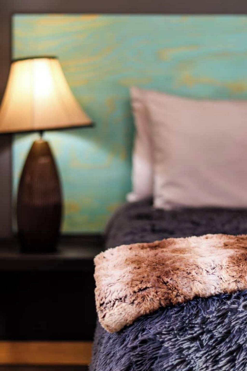 A bed with a fluffy blanket, a lamp on a bedside table, and a wall with a painted design.
