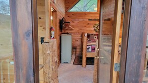 Ein Holzblock-Interieur mit einer kleinen Küchenzeile, einem Bett mit gemusterten Textilien und einem Fenster mit Blick auf Bäume.