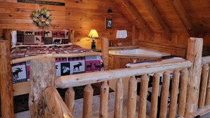 Ein gemütliches Blockhaus-Schlafzimmer mit Bett, Nachttisch, Lampe, kleinem Tisch und einem Fenster mit Blick auf Bäume.
