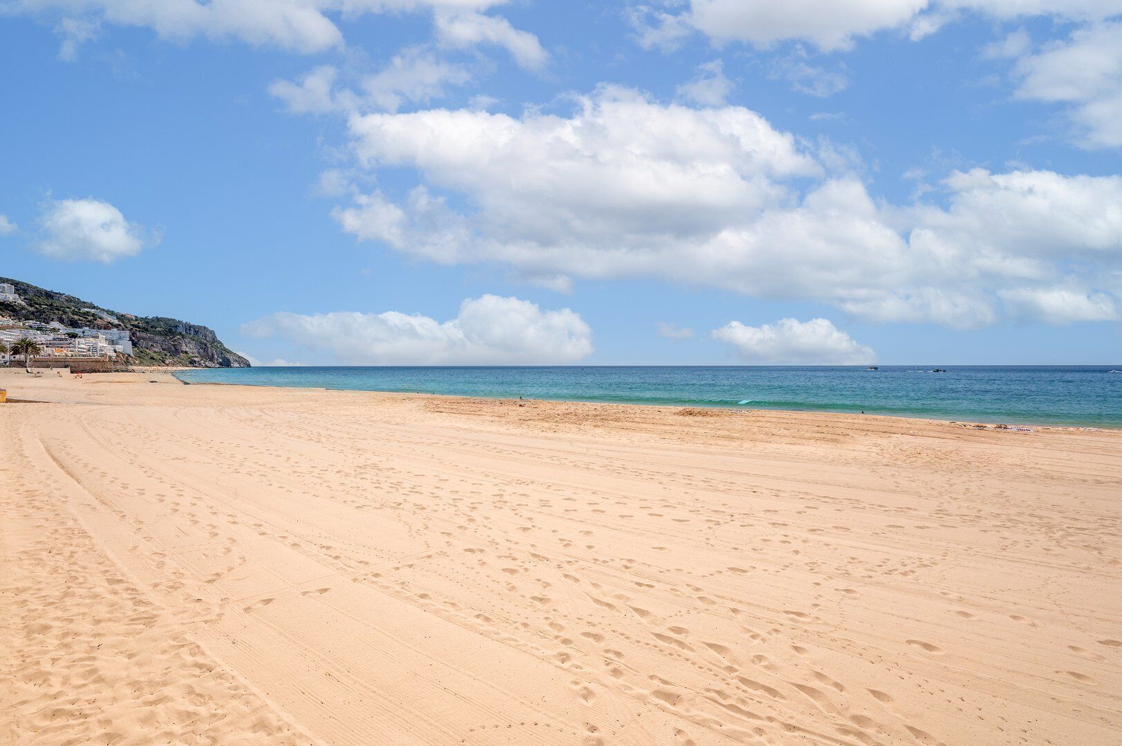 A wide sandy beach with footprints, a clear blue sky, and a calm sea.