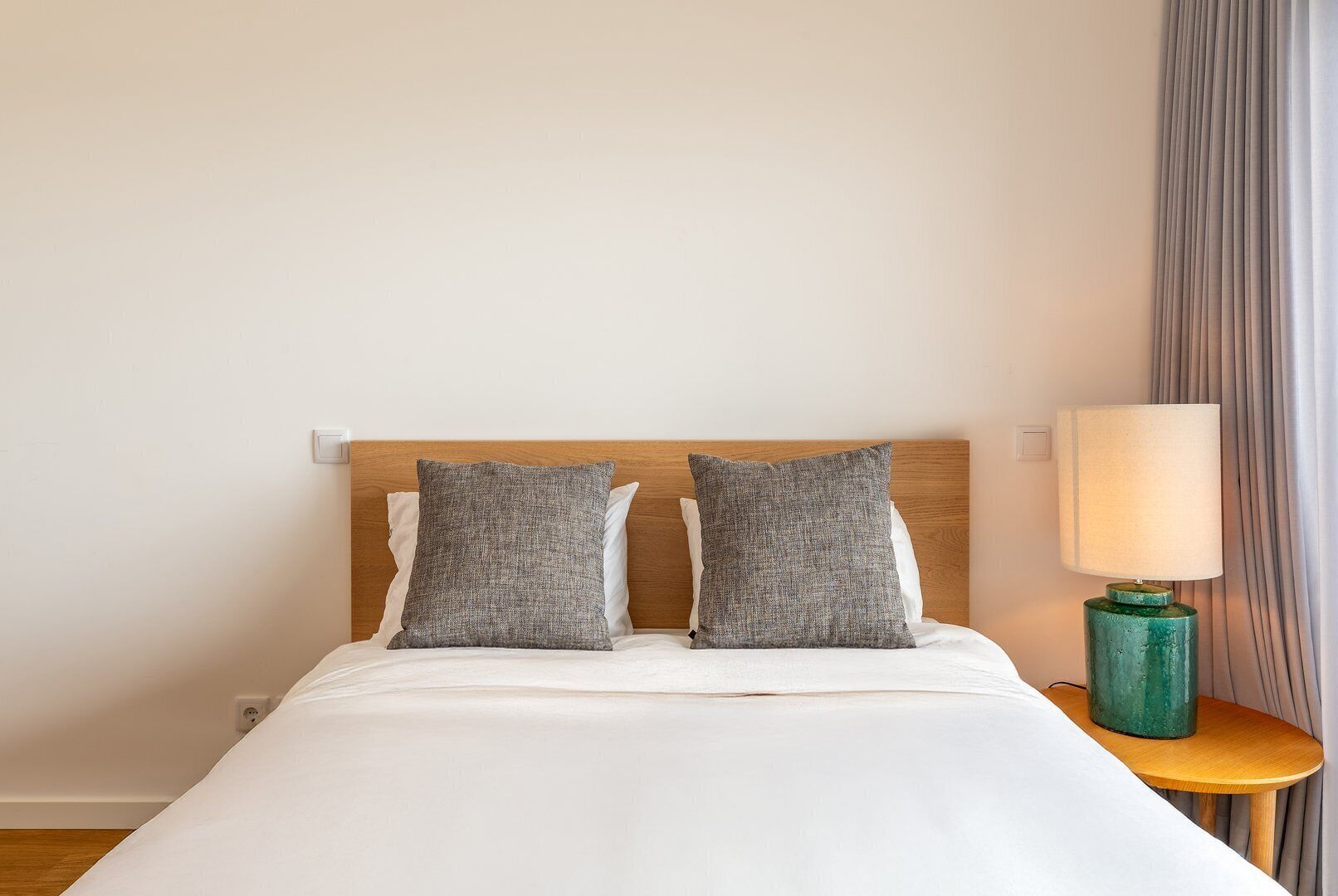 A neatly made bed with two pillows, a wooden headboard, a bedside lamp, and a curtain.