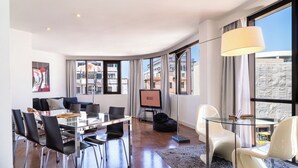 Une chambre d’hôtel moderne dotée d’un coin repas, d’un canapé, d’une télévision et de grandes fenêtres offrant une vue sur la ville.