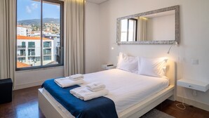 Une chambre d’hôtel avec une grande fenêtre, un lit avec du linge de lit blanc, un élément décoratif bleu sur le lit, un miroir et une table de chevet.