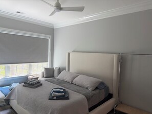 Un dormitorio con cama, ventilador de techo, ventana con persiana y mesita de noche con lámpara.