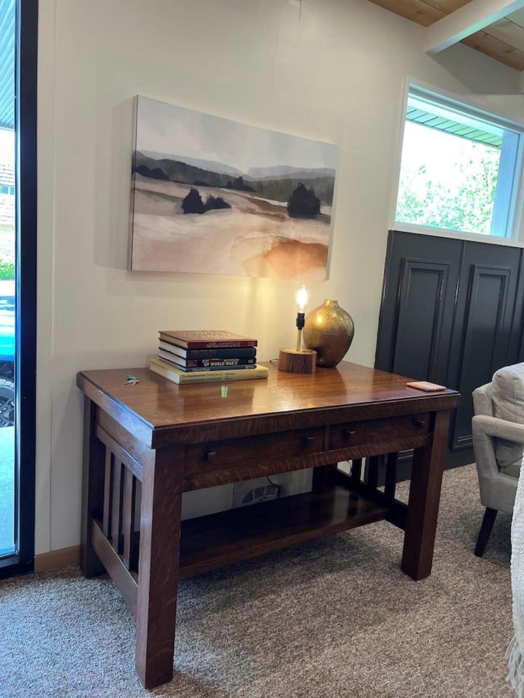 Un escritorio de madera con una lámpara y libros, un cuadro en la pared y vistas al exterior.