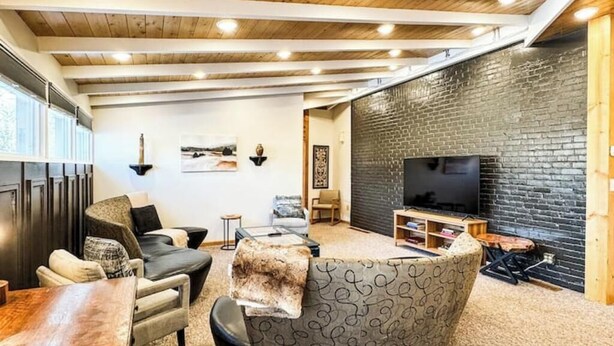 A cozy living room with a wooden ceiling, a round seating area, a flat-screen TV, and a brick accent wall. - MY PLACE! Luxury 2 Bed with piano! (Willmar)