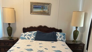 Un dormitorio con una cama cubierta con un colcha azul y blanca con motivos florales, dos lámparas de noche, un cabecero de madera, un cuadro en la pared y una puerta con manija.