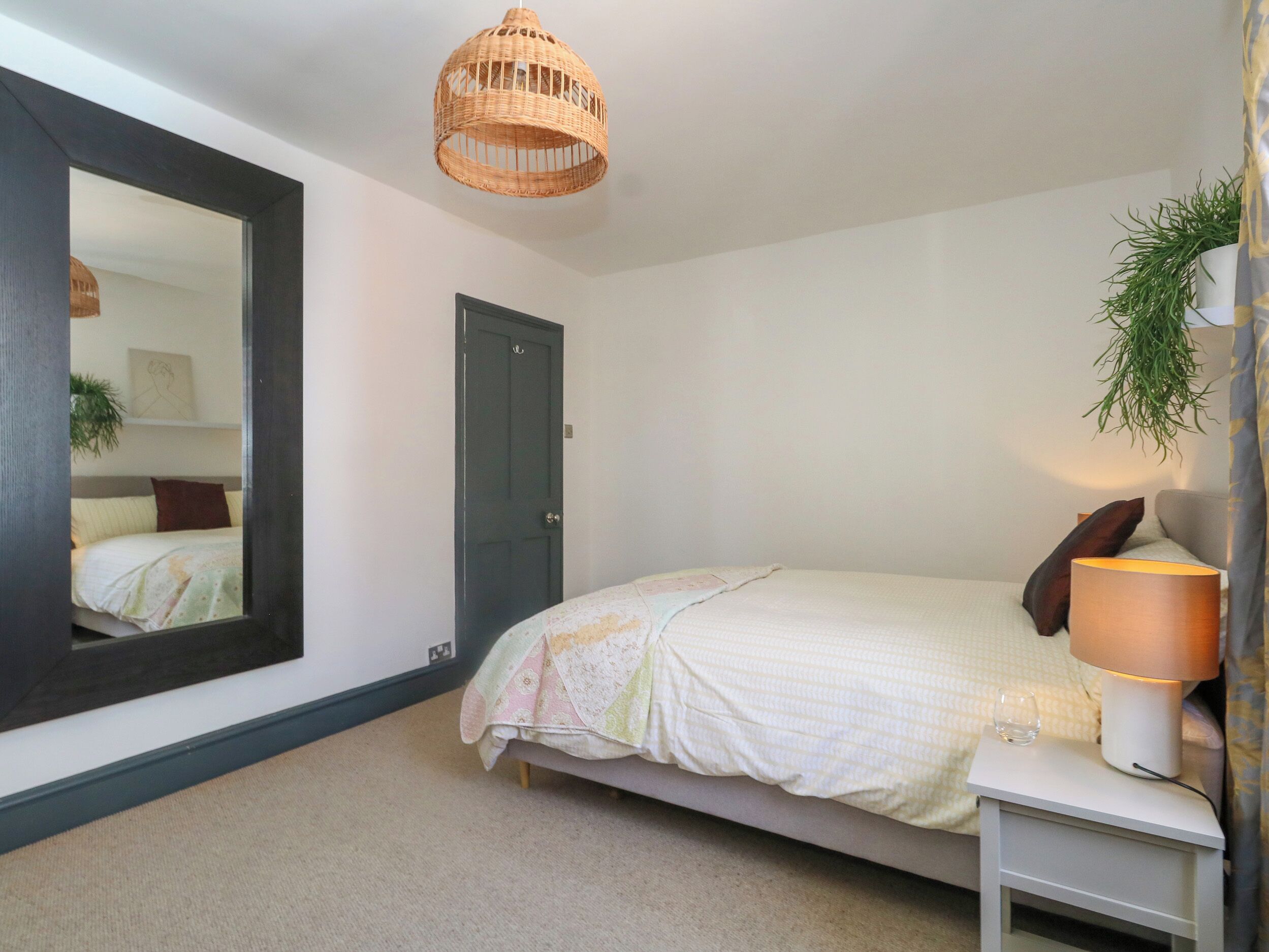 A bedroom with a bed, a bedside table with a lamp, a hanging plant, and a large mirror on the wall.