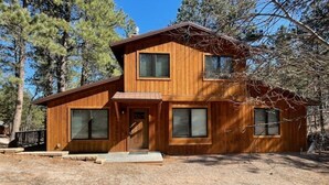 A two-story wooden cabin with a front porch, large windows, and a surrounding forest.
