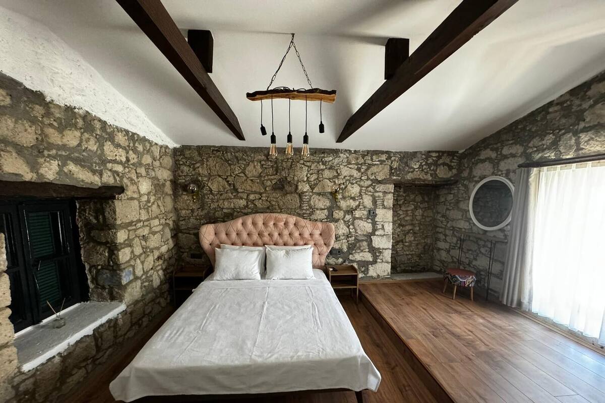A bedroom with a stone wall, a wooden ceiling, a tufted headboard, and a hanging light fixture.